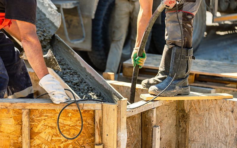 concrete-mixers-workers-are-using-a-hose-to-pour-concrete-into-a-wooden-form-one-worker-stands-on-the-form-while-another-supports-the-hose (1)